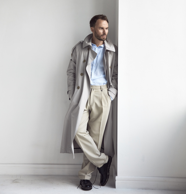 A man wearing a light trench coat standing near a white wall